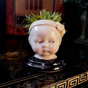 May include: A cream-coloured doll head planter with a cracked finish, holding a green succulent. The planter sits on a black, ornate table. A vintage radio and lamp are in the background. The doll head has a black base.