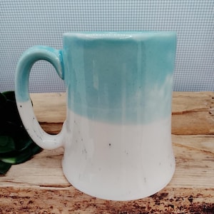 May include: A two-tone ceramic mug, with a turquoise top and white base. The mug features a curved handle and speckled detailing. The mug is displayed on a wooden surface. The mug's height is approximately 12 cm.