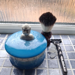 May include: A round blue ceramic shaving soap bowl with a lid featuring a sculpted figure of a person's head, accompanied by a dark brown handled shaving brush and a grey Gillette razor with a multi-blade cartridge head and three small stones.