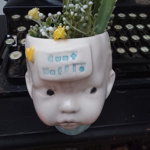 May include: A white ceramic baby-head planter filled with greenery and yellow flowers, with the words "Cunt Waffle" in blue. An antique typewriter is in the background. The planter is a unique decorative item.