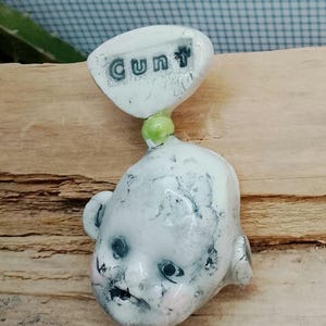 May include: A white ceramic doll's head with the word "cunt" printed on a white oval above it. The doll's head has black eyes, a dark mouth, and pink cheeks. A green bead connects the head to the oval.