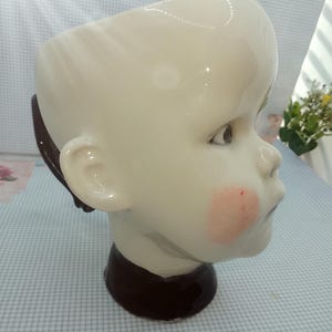 May include: A white ceramic planter shaped like a child's head, with a brown base and accents. The planter has a smooth, glossy finish, with pink blush on the cheeks.