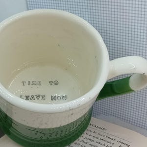 May include: A ceramic mug with a white interior and a dark green exterior base. The interior bottom features the black printed text "TIME TO LEAVE NOW". The mug has a curved handle and a speckled finish on the upper white portion of the exterior.