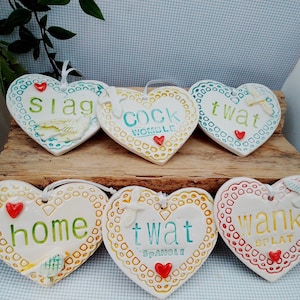 May include: Six white ceramic heart-shaped ornaments with scalloped edges and embossed patterns. Each heart features colourful text including "slag," "cock womble," "twat," "home," "twat spanoli," and "wank splat," accented with small red hearts.