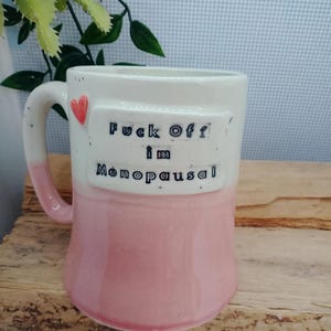 May include: A ceramic mug with a white upper section and a pink lower section. The mug features a small red heart near the handle and a raised rectangular plaque with the black stamped text "Fuck Off i'm Menopausal" on the front surface.