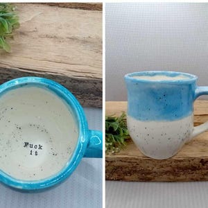 May include: A ceramic mug with a speckled white interior and exterior, featuring a bright blue glazed rim and upper section. The interior base of the mug has the text "Fuck it" stamped in black lettering. The mug has a curved handle and a rounded body.