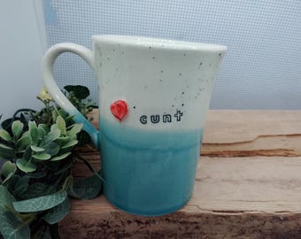 May include: A ceramic mug featuring a two-tone design with a speckled off-white top and a solid teal bottom. The mug includes a small, raised red heart-shaped accent and the word "cunt" printed in black lowercase letters on the front side.