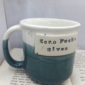 May include: A ceramic mug with a two-tone design featuring a white upper section and a dark teal lower section. A rectangular plaque is attached to the front with the text "Zero Fucks given" in black lettering. The white portion has small dark speckles.