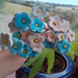 May include: A bouquet of ceramic flowers in a variety of colors, including light blue, pink, and white, with yellow centers. The flowers are arranged in a cluster, with green leaves and a blue pot in the background.