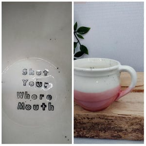 May include: White ceramic mug with a pink ombre design and a curved handle. The mug has the text "Shut Your Whore Mouth" inside. The mug is on a wooden surface.