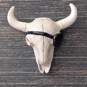 May include: White ceramic bull skull with a black eye patch. The skull is cracked and has a distressed look.