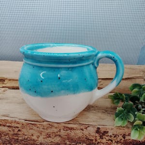 May include: A white ceramic mug with a blue glaze and speckles. The mug has a rounded body and a handle.