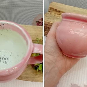 May include: A pink ceramic mug with a rounded body and a handle. The interior is white with black speckles and the words "Sugar Tits" are visible. The mug is on a wooden surface.