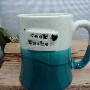 May include: A ceramic mug with a white top and teal bottom. The mug has a handle and the words "Cock Sucker" are printed on the front, along with a black heart.