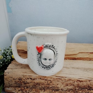 May include: Speckled white ceramic mug with a framed ghostly face and a red heart. The mug has a curved handle and is set on a wooden surface. A unique, handcrafted item for tea or coffee.