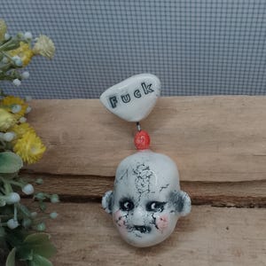 May include: A ceramic pendant featuring a white, cracked-finish doll head with dark eye makeup and pink cheeks, connected by a small red bead to a white, heart-shaped charm with the word "Fuck" printed in black, sans-serif capital letters.