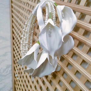 May include: A cluster of white calla lilies with pearl accents and a pearl strand, tied with a white ribbon. The flowers are set against a light brown wooden lattice background, creating a delicate and elegant display.