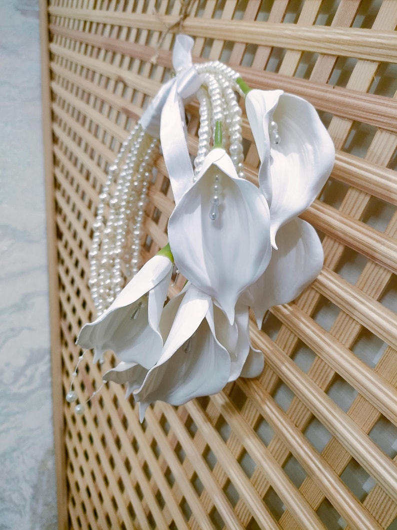 May include: A cluster of white calla lilies with pearl accents and a pearl strand, tied with a white ribbon. The flowers are set against a light brown wooden lattice background, creating a delicate and elegant display.