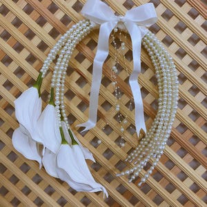 May include: A decorative wreath featuring strands of faux pearls, white calla lilies, and a white satin bow. The wreath is designed with multiple strands of pearls and white ribbons, creating a cascading effect. The calla lilies have green stems and white petals.