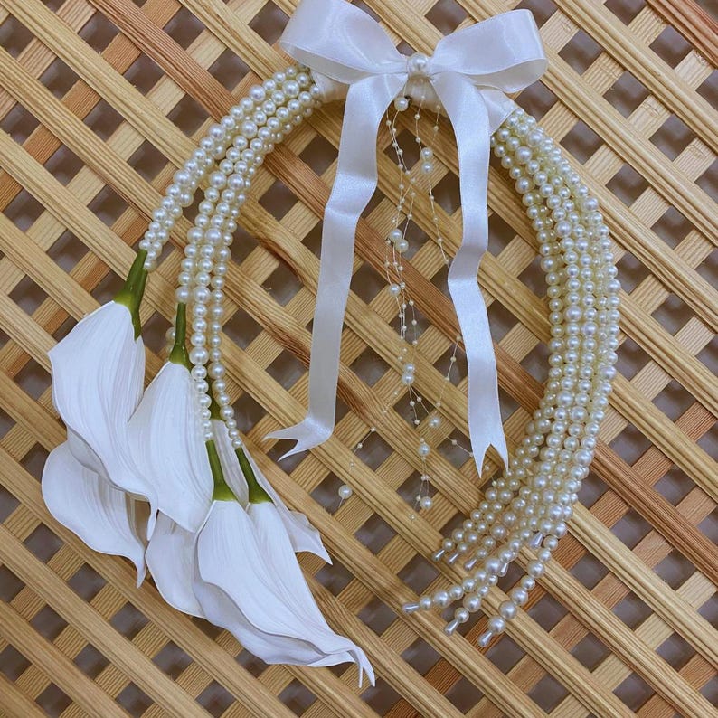 May include: A decorative wreath featuring strands of faux pearls, white calla lilies, and a white satin bow. The wreath is designed with multiple strands of pearls and white ribbons, creating a cascading effect. The calla lilies have green stems and white petals.