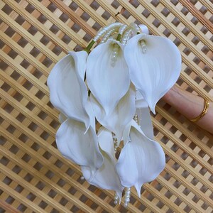 May include: A bouquet of white calla lilies with pearl accents. The flowers are arranged with a satin ribbon and pearl strands. The background is a light brown lattice.