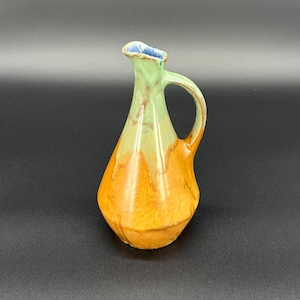 Peut inclure: Un petit pichet en céramique fait à la main avec une poignée. Le pichet présente un dégradé de couleurs, passant d'un vert clair en haut à une base orange doré. Le bord intérieur est bleu. Le pichet est probablement utilisé pour servir des liquides.