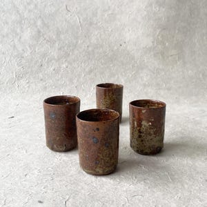 Peut inclure: Quatre petites tasses en céramique brune avec un glaçage moucheté. Les tasses sont disposées en rangée sur une surface blanche.