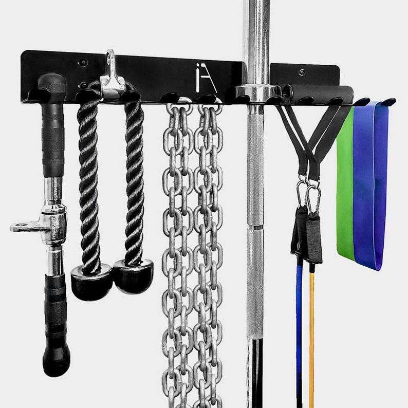 Puede incluir: Un estante de almacenamiento de metal negro montado en la pared con ganchos para colgar equipos de gimnasio. El estante tiene una barra de pesas, dos asas de cuerda, una cadena, dos bandas de resistencia y una barra de levantamiento de pesas.