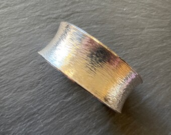 Könnte beinhalten: Ein silberfarbenes Armreif-Armband mit strukturierter, gehammerter Oberfläche. Das Armband hat eine gebogene Form und ein breites Band. Die Oberfläche reflektiert das Licht und erzeugt Glanzlichter und Schatten. Das Schmuckstück wird auf einer dunkelgrauen Oberfläche präsentiert.