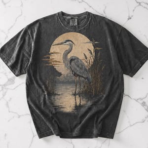 May include: Dark grey t-shirt with a heron illustration set against a sunset scene. The design includes reeds and a tranquil landscape. The shirt has a vintage, washed appearance. The artwork evokes a nature theme, suitable for casual wear.
