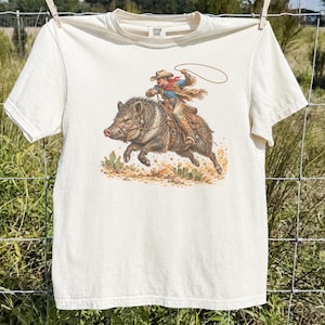 May include: A cream-coloured short-sleeve t-shirt featuring a graphic print of a person wearing a cowboy hat and red scarf, riding a large wild boar while holding a lasso. The illustration includes desert elements like small cacti and sandy ground.