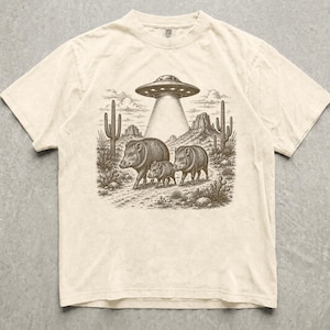 Könnte beinhalten: Ein cremefarbenes Kurzarm-T-Shirt mit einem dunkelbraunen Grafikdruck von drei Pekaris, die durch eine Wüstenlandschaft mit Kakteen und Felsformationen unter einer fliegenden Untertasse laufen, die einen Lichtstrahl ausstrahlt.