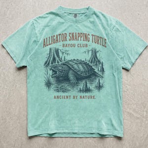 Puede incluir: Una camiseta de manga corta de color verde claro con un gráfico en verde oscuro de una tortuga caimán en un pantano. El texto dice ALLIGATOR SNAPPING TURTLE - BAYOU CLUB - ANCIENT BY NATURE. La camiseta tiene cuello redondo y un acabado desgastado.