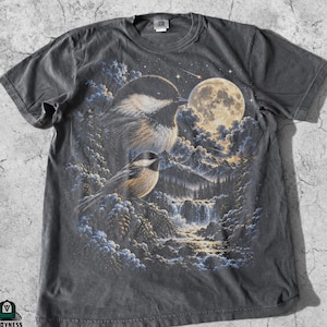 May include: A charcoal gray short-sleeve t-shirt featuring a graphic print of two chickadees in a mountain landscape with a full moon, clouds, and a waterfall. The Vroyness Apparel logo is printed in the bottom left corner of the shirt.