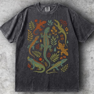Op de afbeelding: Een antracietgrijs T-shirt met korte mouwen en een mineral wash-afwerking. De voorkant heeft een grafische print van vijf hagedissen in tinten blauwgroen, oranje en geel, afgewisseld met botanische illustraties van bladeren en bloemen.
