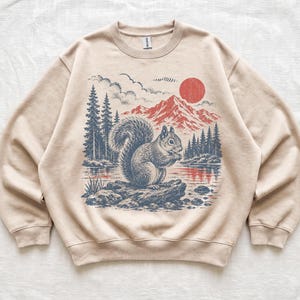 May include: A beige crewneck sweatshirt featuring a graphic illustration of a squirrel sitting on a log by a lake. The scene includes dark blue pine trees, red mountains, and a red sun in the sky. The design is centred on the front of the garment.