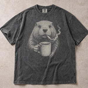 May include: A charcoal grey, acid-washed short-sleeve t-shirt featuring a detailed graphic of a groundhog holding a coffee mug and smoking a cigar. The groundhog is rendered in shades of grey and white, with smoke curling upward from the cigar.