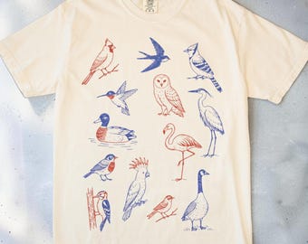 Puede incluir: Una camiseta de manga corta de color crema con un patrón repetitivo de varias especies de aves impresas en tinta roja y azul. El gráfico incluye ilustraciones de un cardenal, una golondrina, un arrendajo azul, un colibrí, un búho y otras aves.