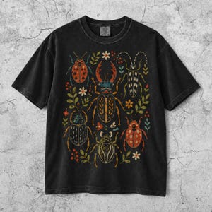 Könnte beinhalten: Ein schwarzes T-Shirt mit kurzen Ärmeln und einem Grafikdruck von fünf verschiedenen Käfern in Rot-, Orange- und Gelbtönen, durchsetzt mit kleinen grünen Blättern und weißen Blumen. Das T-Shirt hat einen Rundhalsausschnitt und eine verwaschene Optik.