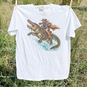 Op de afbeelding: Een wit t-shirt met korte mouwen en een grafische print van een cowboy met een hoed en een lange jas die op een grote alligator rijdt. De alligator is afgebeeld met een open bek terwijl hij door het water spat.
