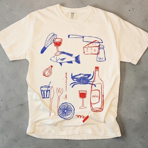 Puede incluir: Camiseta de color crema con una ilustración azul y roja de elementos culinarios. El diseño incluye un pescado, copas de vino, una botella, queso y otros utensilios de cocina. Las palabras "la cuisine" están escritas en la camiseta.