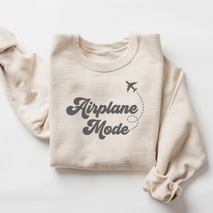 K&ouml;nnte beinhalten: Beiges Sweatshirt mit dem Schriftzug "Airplane Mode" in Retro-Schrift, begleitet von einer kleinen Flugzeuggrafik und einer gepunkteten Flugbahn. Die &Auml;rmel sind hochgekrempelt. Das Sweatshirt ist aus weichem Material.