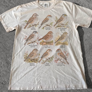 May include: A cream-colored short-sleeve t-shirt featuring a graphic print of nine different sparrow species with their names printed below each bird: song, chipping, white-throated, white-crowned, fox, savannah, Lincoln's, field, and American tree sparrow.