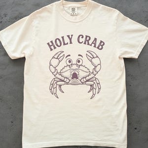 May include: Cream-colored t-shirt featuring a cartoon crab illustration with the text "HOLY CRAB" above it. The crab has wide eyes and open claws. The shirt is made of a soft material.