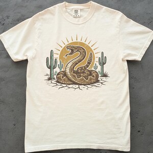 May include: Cream-coloured t-shirt with a desert-themed graphic. The design features a coiled rattlesnake, cacti, and a sunburst. The illustration uses shades of brown, tan, and green, creating a vintage aesthetic.