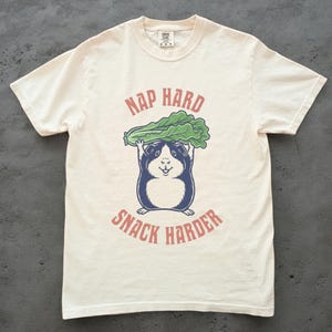 May include: Cream-colored t-shirt featuring a cartoon guinea pig holding a green lettuce leaf above its head. The text "NAP HARD" is arched above the lettuce, and "SNACK HARDER" is arched below. The guinea pig is blue and white.