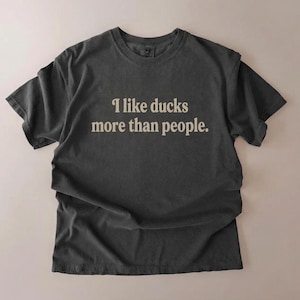 May include: Dark gray t-shirt with the text "I like ducks more than people" in a light beige color. The shirt is made of a soft material and has a crew neck.