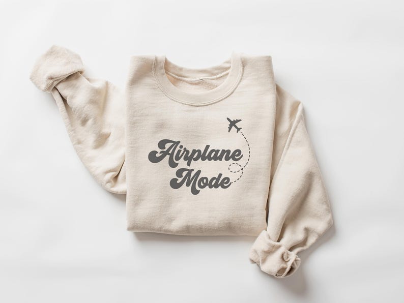 K&ouml;nnte beinhalten: Beiges Sweatshirt mit dem Schriftzug "Airplane Mode" in Retro-Schrift, begleitet von einer kleinen Flugzeuggrafik und einer gepunkteten Flugbahn. Die &Auml;rmel sind hochgekrempelt. Das Sweatshirt ist aus weichem Material.