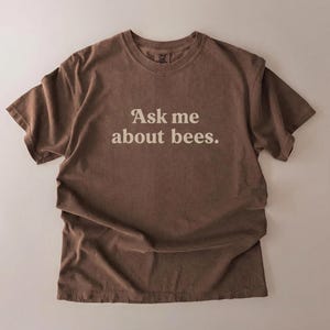 May include: A brown t-shirt with the text "Ask me about bees" in a white, sans-serif font. The shirt is made of a soft material and has a relaxed fit. The t-shirt is laid flat on a neutral background.