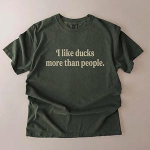 May include: Olive green t-shirt with the text "I like ducks more than people" in a white, sans-serif font. The shirt has short sleeves and a crew neck. The fabric appears to be soft and slightly faded.